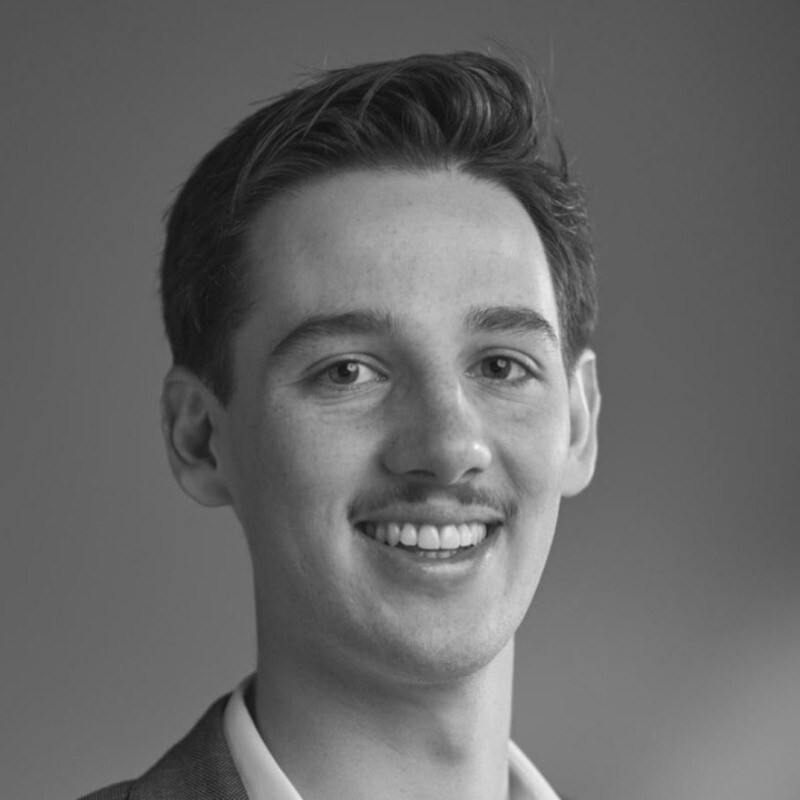 Portrait of a young man smiling with short, styled hair and a faint mustache, wearing a collared shirt and jacket.