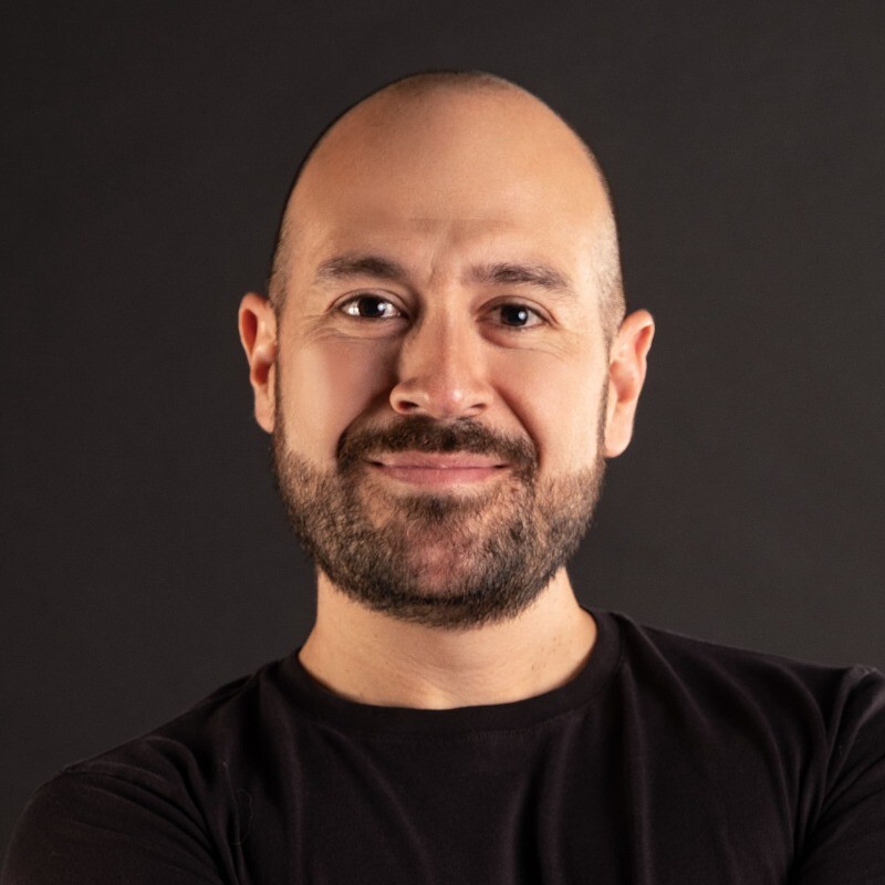 Smiling bald man with a beard wearing a black shirt against a dark background.