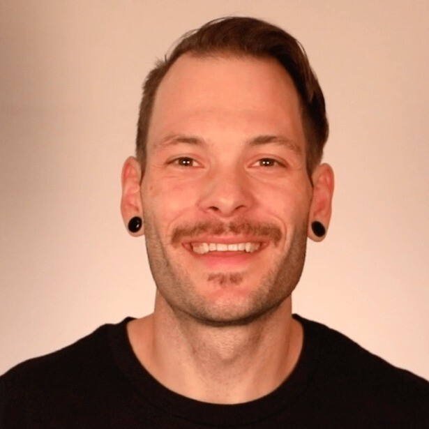 Smiling man with short brown hair, trimmed mustache, and black ear gauges wearing a black shirt against a beige background.