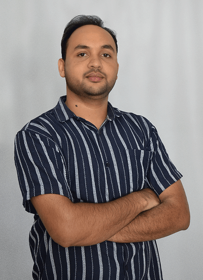 Man with short black hair and beard wearing a black and white striped short-sleeve shirt with arms crossed, standing against a light grey background.